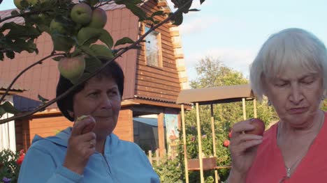 Two-senior-women-trying-apples-in-orchard-near-big-house