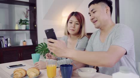 Attractive-young-Asian-couple-distracted-at-table-with-newspaper-and-cell-phone-while-eating-breakfast.-Excited-young-Asian-couple-surprised-by-unbelievably-good-news,-happy-family-amazed-by-internet.