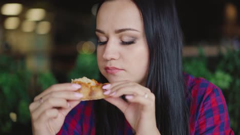 beautiful-brunette-in-a-plaid-shirt-is-eating-pizza-with-enjoy-in-the-cafe-on-the-background-of-the-interior