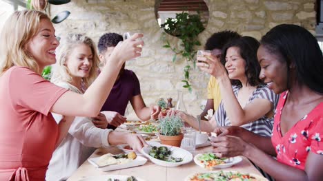 Eine-multi-ethnische-Gruppe-von-gemischten-Altersgruppen-Erwachsene-Freunde-Essen-gemeinsam-am-Tisch-in-einem-Restaurant-erheben-ihr-Glas-in-einem-Toast,-Seitenansicht