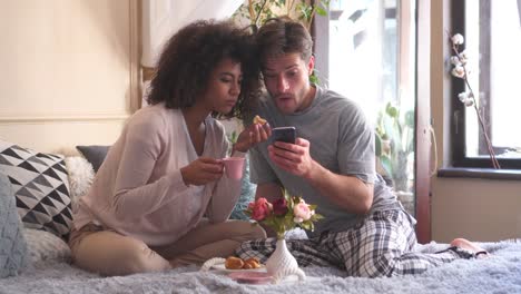 Couple-having-breakfast-while-using-mobile-phone-in-bed