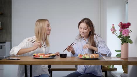 Two-lovely-young-girls-eating-sushi-rolls-and-talking,-friends-together-in-kitchen