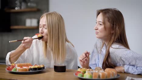 Dos-hermosas-chicas-comen-comida-japonesa-en-casa.-Rollos-en-una-placa-son-diferentes,-muy-sabrosos