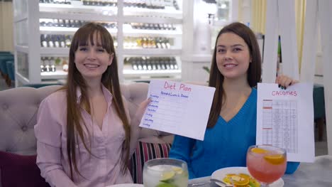 good-nutrition,-portrait-of-happy-girls-holding-in-hands-diet-plan-and-calories-list-and-look-on-camera-in-cafe