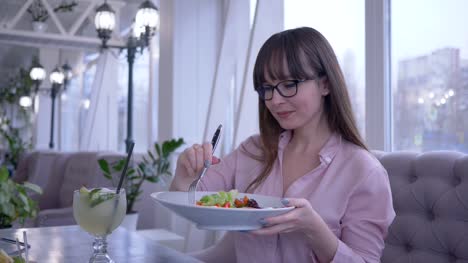 Ernährung,-fröhliches-junges-Mädchen-in-Gläsern-sitzen-im-Café-mit-einer-Gabel-in-der-hand-griechischen-Salat-essen-und-schaut-in-die-Kamera-zu-starten