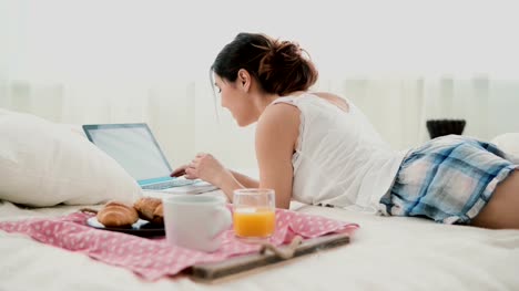 Brunette-girl-lying-on-bed-in-morning-and-chatting,-using-laptop-for-that.-Young-woman-typing-on-keyboard-and-laughing