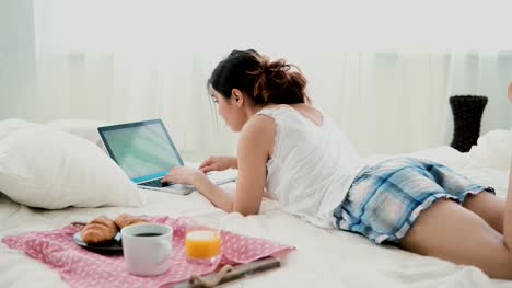 Joven-usando-laptop-durante-el-desayuno-de-mentira-en-cama-blanca-en-casa.-Chica-morena-escribiendo-en-la-computadora-de-la-pc