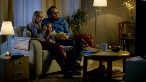 Totale-von-Vater,-Mutter-und-Mädchen-vor-dem-Fernseher.-Sie-sitzen-auf-einem-Sofa-in-ihrem-gemütlichen-Wohnzimmer-und-Popcorn-essen.-Es-ist-Abend.