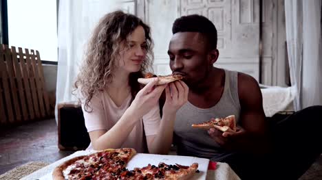 Multiracial-couple-have-fun-during-the-meal.-Woman-feed-the-man-a-slice-of-pizza.-Male-and-female-eating-fast-food