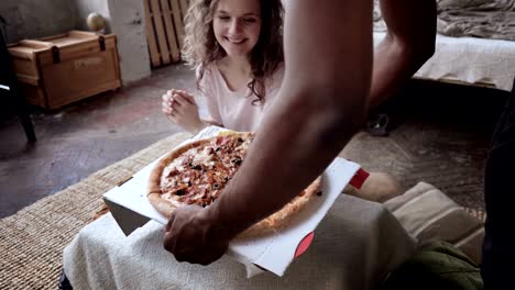 Hombre-africano-trae-pizza-a-una-mujer-caucásica-de-feliz.-Una-pareja-multirracial-disfrutando-de-buena-comida,-sonriendo,-riendo