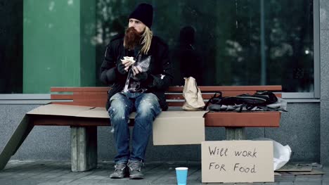 Obdachlos-und-arbeitsloser-amerikanischer-Mann-mit-Pappschild-Sandwich-Essen-auf-Bank-am-Stadtstraße-wegen-Krise-der-Einwanderer-in-den-usa