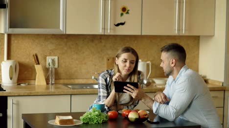 Atractiva-pareja-utilizando-tablet-computadora-fo-social-medios-de-comunicación-y-tener-el-desayuno-en-la-cocina