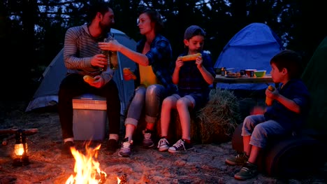 Eltern-trinken-Bier-und-Essen-auf-frischen-gelben-Mais-aus-Feuer-während-der-Sommerferien,-Abendessen-in-der-Nähe-von-Flammen