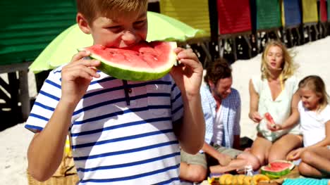 Boy-eating-watermelon-while-family-sitting-in-background-at-beach