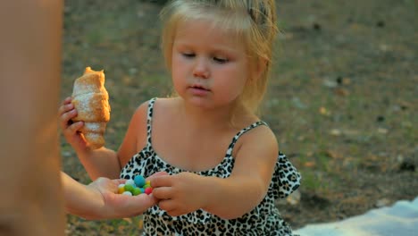 Little-girl-eats-a-croissant-and-tries-a-colorful-candies-from-female's-hand