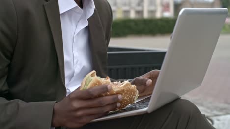 Closeup-del-empresario-Burger-de-mascar-y-el-uso-de-ordenador-portátil-al-aire-libre