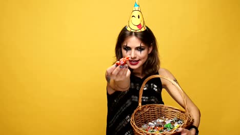 Tricky-lady-with-halloween-make-up-and-holiday-cap-teasing-with-candies-and-laughing-isolated-over-yellow