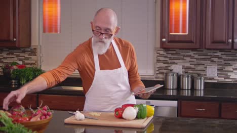 Old-chef-reaches-out-to-grab-some-vegetables-while-he-uses-his-tablet