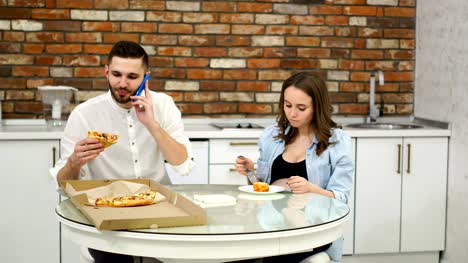 Mann-und-schwangere-Frau-Essen-Pizza-zu-Hause-in-ihrer-Küche.-Mann-am-Telefon-zu-sprechen,-während-des-Abendessens