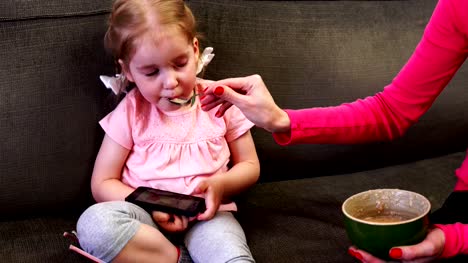 Ein-kleines-Mädchen-schaut-auf-ihr-Smartphone,-während-ihre-Mutter-ihr-einen-Löffel-füttert