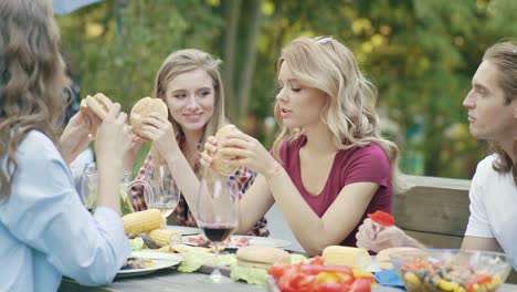 Friends-Eating-Burgers-Sitting-At-Dinner-Table-At-Outdoor-Party