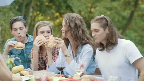 Freunde-Essen-Burger-sitzen-am-Esstisch-auf-Outdoor-Party