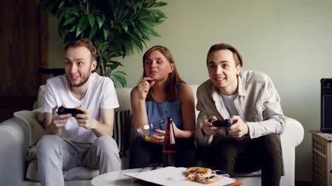 Two-male-friends-are-playing-video-games-pressing-buttons-on-joystick-while-pretty-blond-girl-is-watching-game-and-eating-snacks.-Friendship-and-technology-concept.