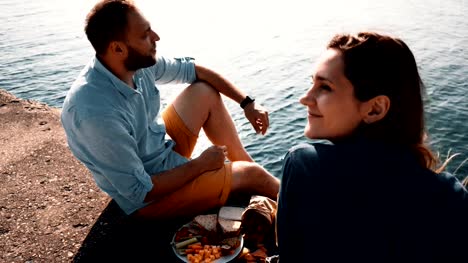 Young-happy-couple-sitting-on-the-shore-of-a-sea-and-eating.-Man-and-woman-having-picnic-near-water-in-the-morning
