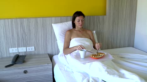 Una-mujer-es-comer-waffles-con-un-cuchillo-y-el-tenedor-en-la-cama-en-una-habitación-de-hotel