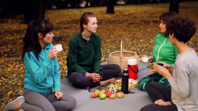 Schöne-Sportlerinnen-sind-entspannend-nach-outdoor-Training-mit-Picknick-auf-Matten-Snacks-zu-essen-und-trinken-Tee.-Mädchen-sind-reden-und-lachen-genießen-Natur.