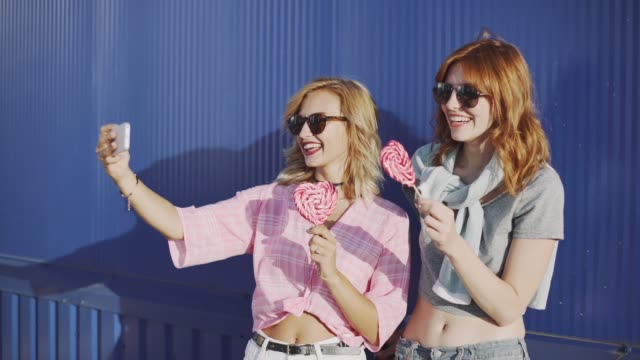 Dos-mujeres-jóvenes-atractivas-en-gafas-de-sol,-hablar,-comer-Chupa-Chups-y-tomar-un-selfie-en-calle