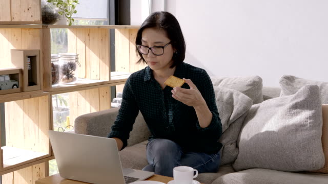 Schöne-junge-Frau,-die-Asiaten-mit-Computer-Laptop-arbeiten-und-Essen,-Snacks,-Kaffee-trinken,-beim-Sitzen-auf-dem-Sofa-zu-Hause.-Arbeit-zu-Hause-Konzept