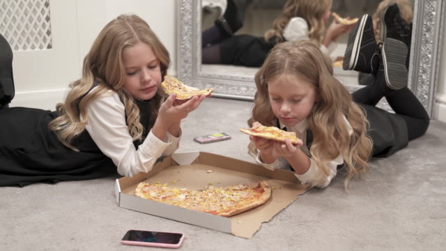 Zwei-ziemlich-blonde-Mädchen-liegen-auf-dem-Boden-essen-Pizza.-Es-ist-eine-geöffnete-Box-mit-Pizza-vor-ihnen.-Sie-blicken-auf-den-Handy-Bildschirm.