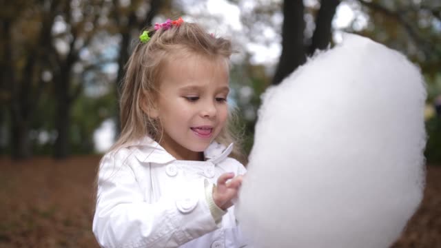 Little-blonde-girl-is-eating-sweet-cotton-candy-in-the-city-park.-Beautiful-little-girl-eating-candy-floss.-Child-eating-cotton-candy-green-forest-at-the-backgorund.-4k.-Slow-Motion