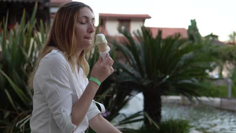 Portrait-of-beautiful-positive-young-woman-eating-ice-cream-on-background-of-palm-trees