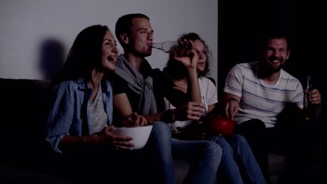 Friends-of-four-sit-on-couch,-watch-funny-film-on-TV.-Caucasian-young-group-sit-on-the-couch,-drinks-and-snacks.-Movie-night