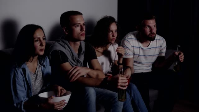 Four-friends-eating-popcorn,-drinking-beer-watch-horror-movie-together-and-are-very-captivated-and-scared,-sit-still.-Movie-night