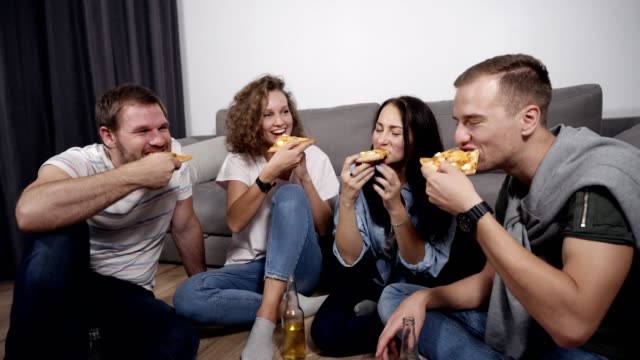 Group-of-four-friends-having-fun,-ordered-large-pizza-and-eating,-laughing-and-talking,-sitting-on-floor-in-grey-and-white-coloured-loft-room.-Drinking-and-spending-time-together