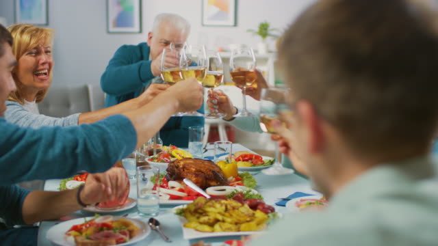 Große-Familie-und-Freunde-feiern-zu-Hause,-vielfältige-Gruppe-von-Menschen-versammelten-sich-am-Tisch-anstoßen-in-einem-Toast.-Leute-Essen,-trinken-und-Spaß-haben.-Tagsüber-Festlichkeit-im-Wohnzimmer.