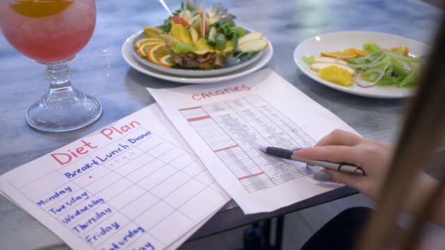 Ernährungsberaterin-Frau-auf-der-Suche-nach-Kalorien-Liste-und-halten-in-der-hand-sitzen-am-Tisch-mit-Punsch-und-Früchte-in-Platte-Diät-plan