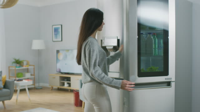 Beautiful-Brunette-Girl-in-White-Jeans-and-Grey-Sweater-Comes-to-the-High-Tech-Fridge-with-a-Glass-Door-and-Takes-a-Green-Apple.-She-goes-to-the-Living-Room.-Flat-has-Modern-Interior.