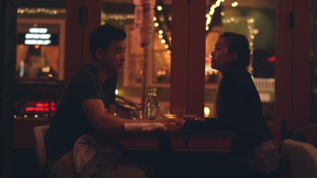 A-young-couple-laughing-on-a-romantic-dinner-date,-with-city-lights-behind-them