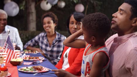 Young-boy-on-dad’s-knee-at-family-barbecue-flexing-muscle