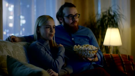 Couple-Watching-TV.-They-Sit-on-a-Sofa-in-Their-Cozy-Living-Room-and-Eat-Popcorn.-It's-Evening.