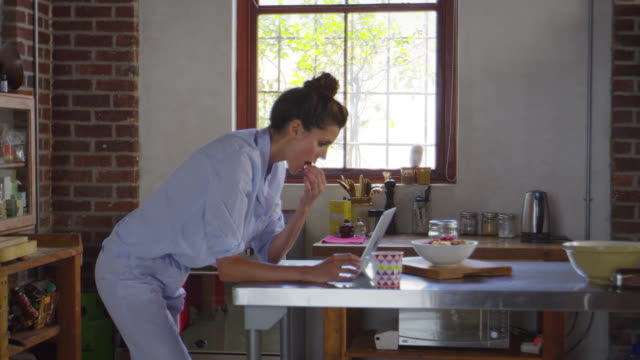 Mujer-joven-en-pijama-usando-computadora-portátil-en-la-cocina,-rodada-en-R3D