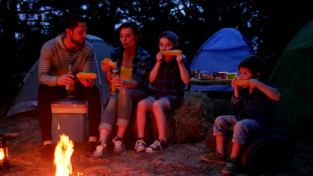 woman-and-male-drink-beer-and-eat-up-fresh-yellow-maize-from-out-fire-during-summer-holidays,-family-enjoying-corn-with-salt