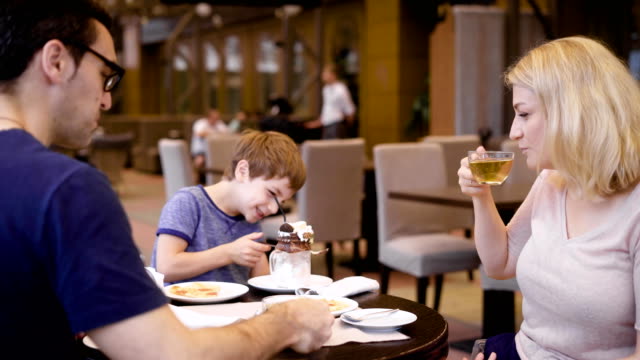 Familia-feliz-con-pequeño-hijo-sentado-en-la-mesa.-Madre-bebiendo-té-y-usar-su-smartphone.-Niño-que-se-divierte-comiendo-postre-cremoso-de-chocolate.-Padres-jóvenes-disfrutando-de-una-cena-en-el-restaurante-con-el-niño