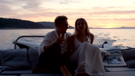 Young-Couple-Hug,-Drink-Champagne-in-the-Stern-of-the-Moving-Yacht.-They-Have-Great-Romantic-Evening.-In-the-Background-Island-with-Small-Village.
