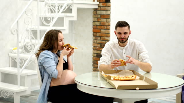 Mann-und-schwangere-Frau-Essen-Pizza-zu-Hause-in-ihrer-Küche.-Lachen-Spaß
