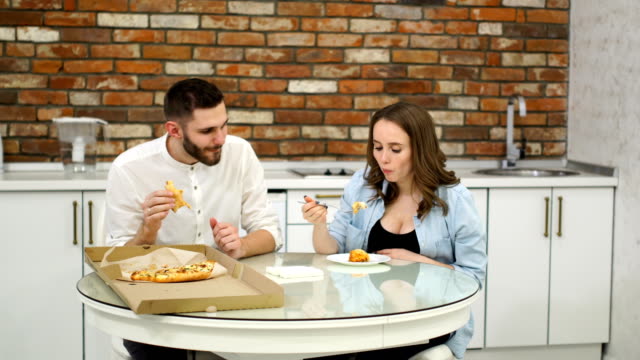 Mann-und-schwangere-Frau-Essen-Pizza-zu-Hause-in-ihrer-Küche.-Schlechte-Ernährung.-Fetthaltigen-Lebensmitteln.-Fettleibigkeit